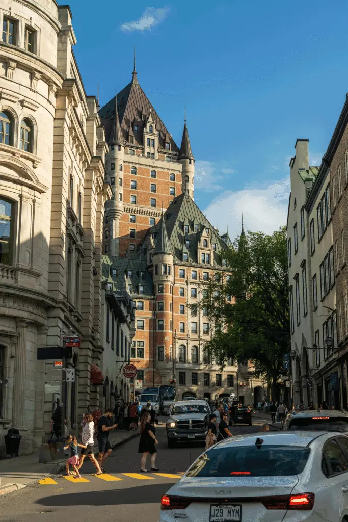 Pedestrians and cars move through Old Quebec, with Château Frontenac rising above the historic buildings under a blue sky.